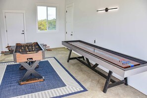 A room with a foosball table, a shuffleboard table, and a window with a view of greenery.