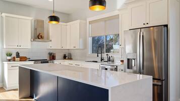 Une cuisine moderne avec des meubles blancs, un grand îlot central, des appareils en acier inoxydable et des luminaires suspendus.