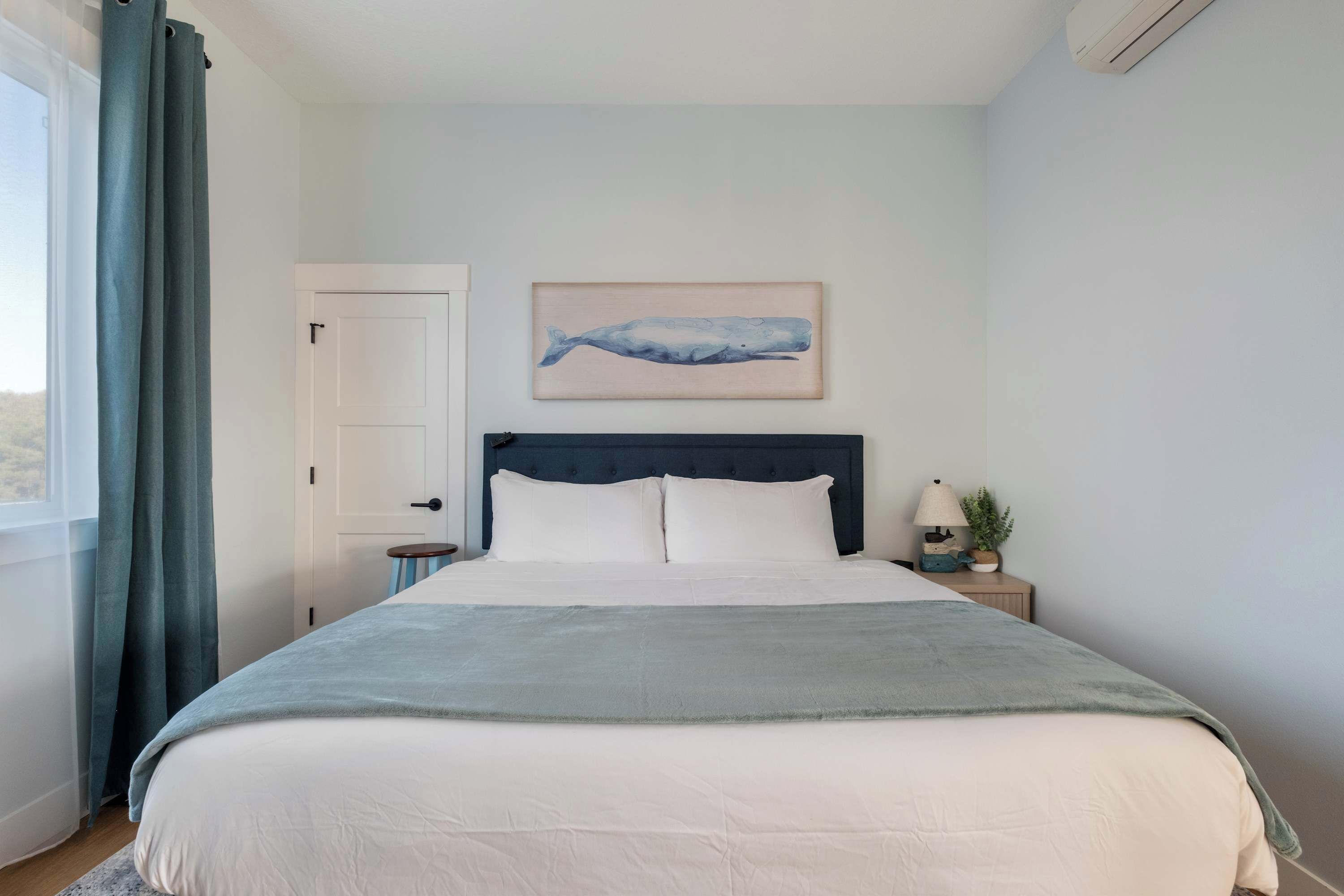 A bedroom with a bed, a nightstand, a wall-mounted air conditioner, a door, and a window with curtains.