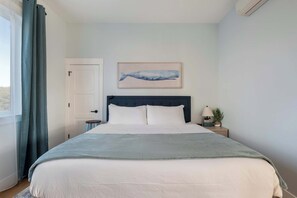 Een slaapkamer met een bed, nachtkastje, een wandairconditioner, een deur en een raam met gordijnen.