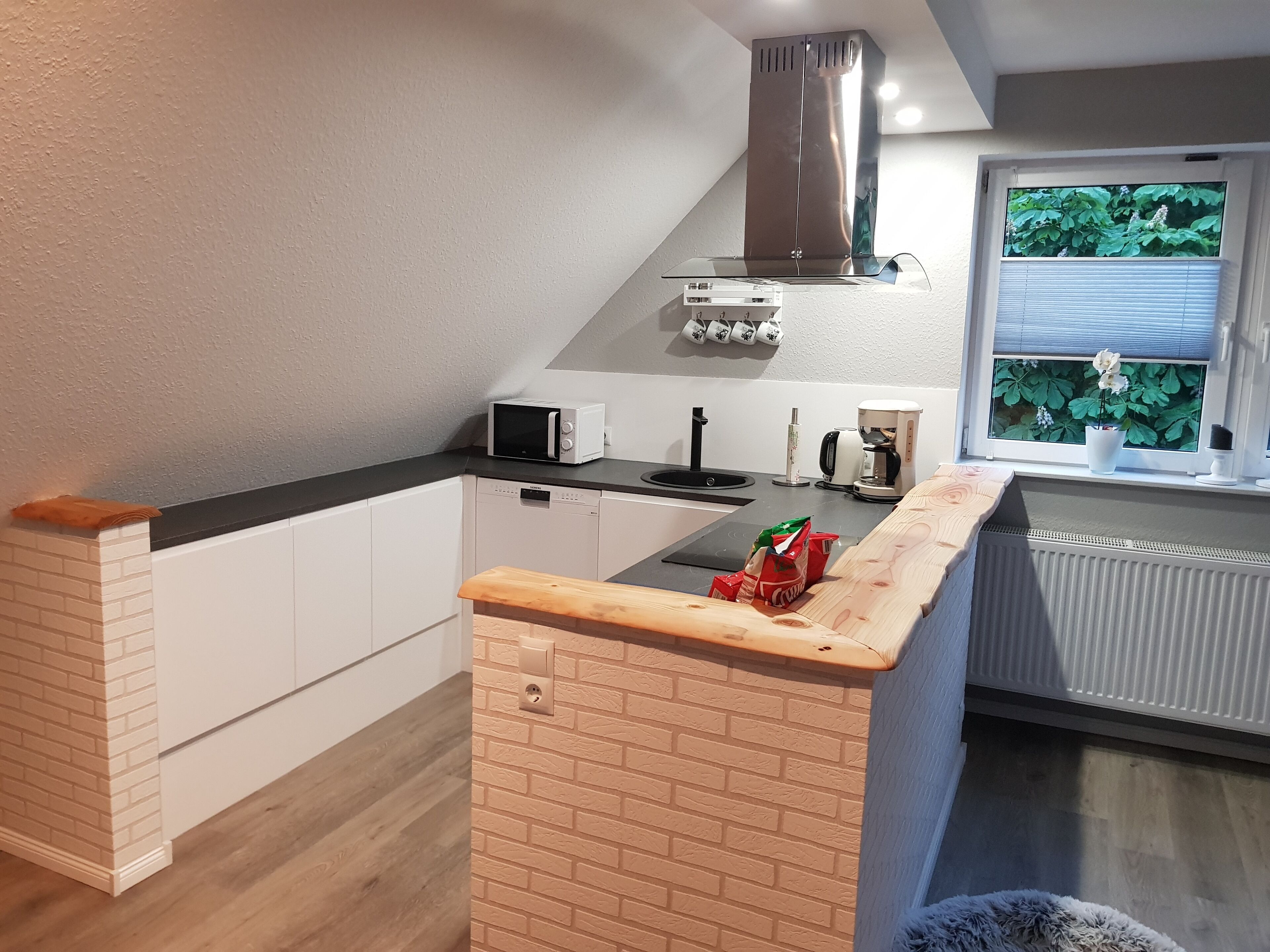 A modern kitchen with white cabinets, a black countertop, a stainless steel range hood, a microwave, a coffee maker, and a window with a view of greenery.