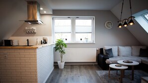 A modern kitchen with a brick backsplash, a white countertop, and a central island. There is a potted plant, a white radiator, and a window with blinds. A cozy seating area with a sofa and two round tables is visible. - Cozy vacation apartment in East Frisia - families & dogs welcome! (Hinte)