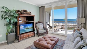 A living room with a television, a brown leather ottoman, a wicker cabinet, a large potted plant, and a view of the ocean through a sliding glass door.