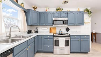 Cocina con armarios azules, electrodomésticos de acero inoxidable, lavavajillas y ventana con vista a árboles.