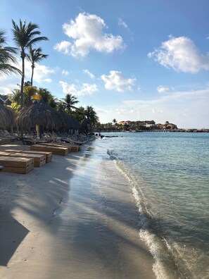 A beach with clear water, palm trees, and thatched umbrellas.