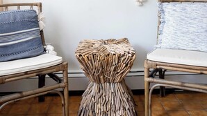 Una habitación con dos sillas de mimbre, un taburete tejido, un colgador de macramé y un adorno de ancla en la pared.