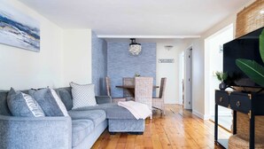 Una sala de estar con un sofá seccional gris, un comedor con sillas de mimbre y un mueble de televisión negro.