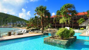 En pool på ett semesterhotell med kristallklart blått vatten, omgiven av palmer och solstolar, med en strand och hotellbyggnader i bakgrunden.