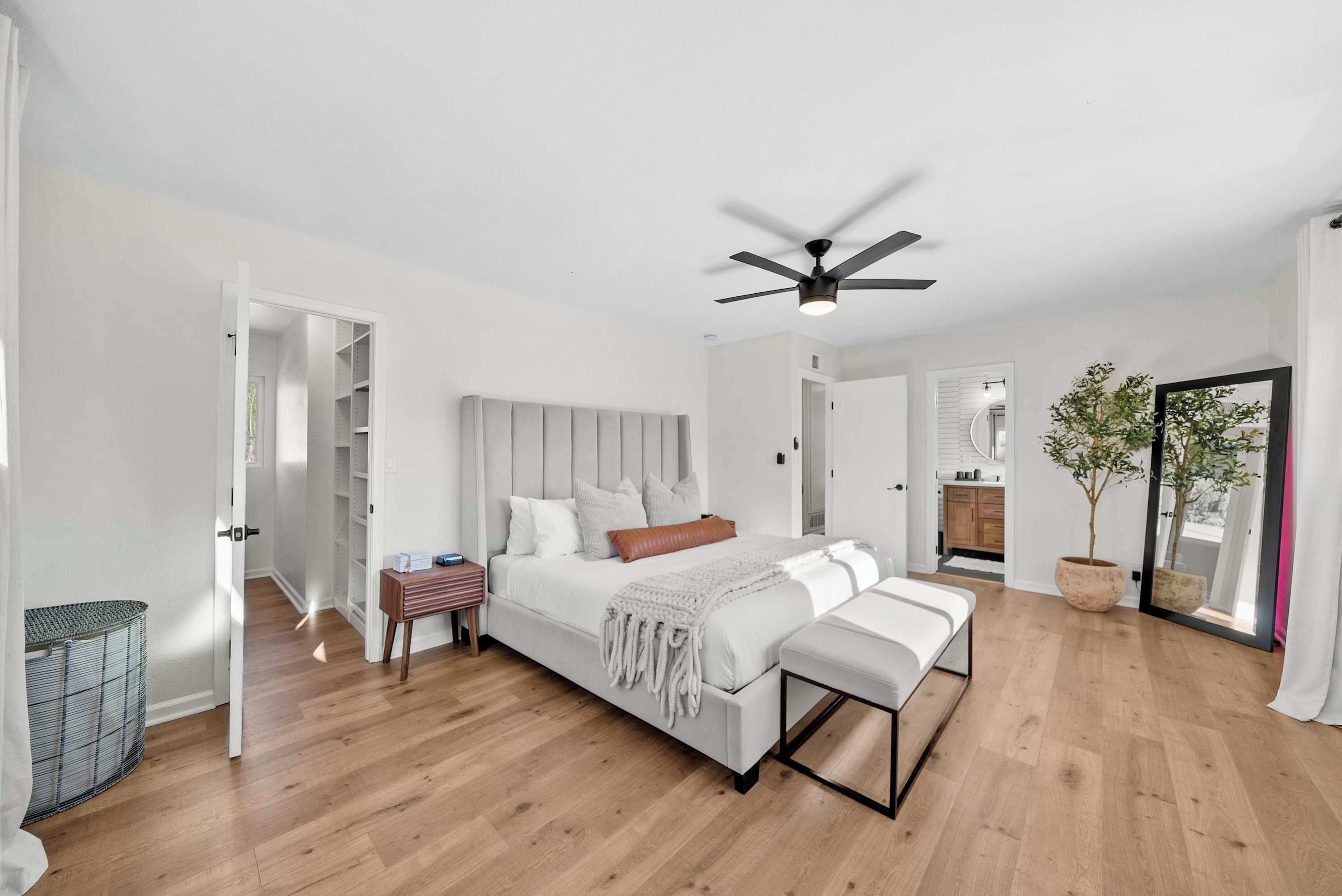 Une chambre moderne avec un grand lit, une table de chevet, un banc, un ventilateur de plafond et une vue sur la cuisine et le balcon.