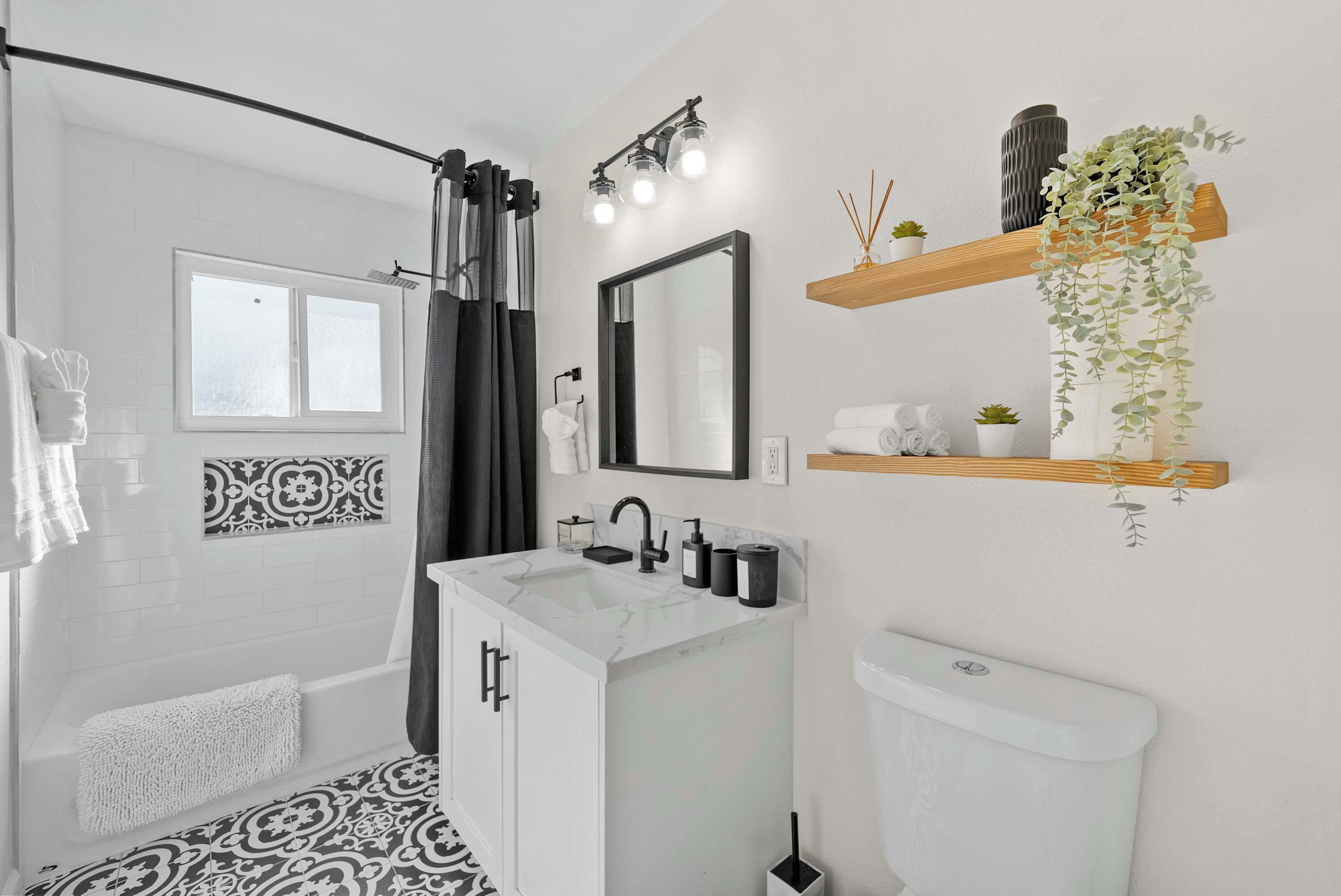 Un baño moderno con ducha, lavabo, inodoro y un espejo. Hay repisas con plantas y artículos de aseo.