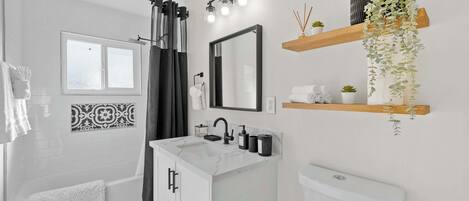 Un baño moderno con ducha, lavabo, inodoro y un espejo. Hay repisas con plantas y artículos de aseo.
