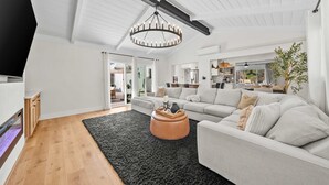 Ein modernes Wohnzimmer mit einem großen Sofa, einem Couchtisch und einer Chandelier. Im Hintergrund ist eine Küche erkennbar.