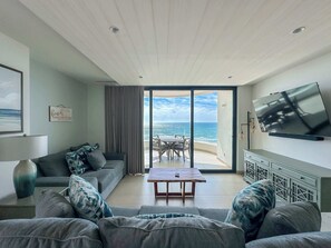 A modern living room with a large TV, a sofa, a coffee table, and a view of the beach.