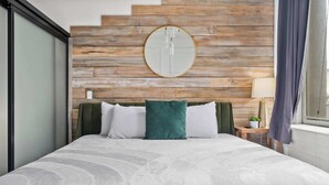 A bed with a white and green patterned bedspread, a wooden headboard with a round mirror, and a small bedside table with a lamp and a plant. - Woolworth Loft - Stay in the Heart of Gaslamp 🌆 (San Diego)