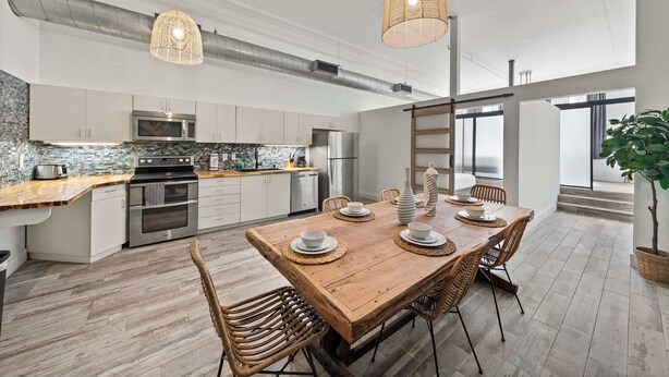 A modern kitchen with stainless steel appliances, a wooden dining table with chairs, and a large window providing natural light. - Woolworth Loft - Stay in the Heart of Gaslamp 🌆 (San Diego)