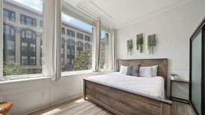 Quarto com uma janela grande, uma cama de madeira e vista para um prédio lá fora.
