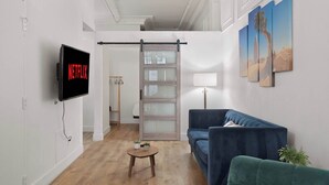 A modern living room with a blue sofa, a small wooden table, and a TV displaying 'NETFLIX'. - Woolworth Loft – Gaslamp 4BR Downtown Pad w/ Tub (San Diego)