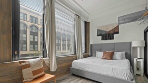 A bedroom with a large bed, wooden floor, and a view of a building through the window. - Woolworth Loft – Gaslamp 4BR Downtown Pad w/ Tub (San Diego)
