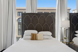 Quarto de hotel com uma cama grande, cabeceira com padrão decorativo, mesa de cabeceira com abajur e vista para edifícios pela janela.