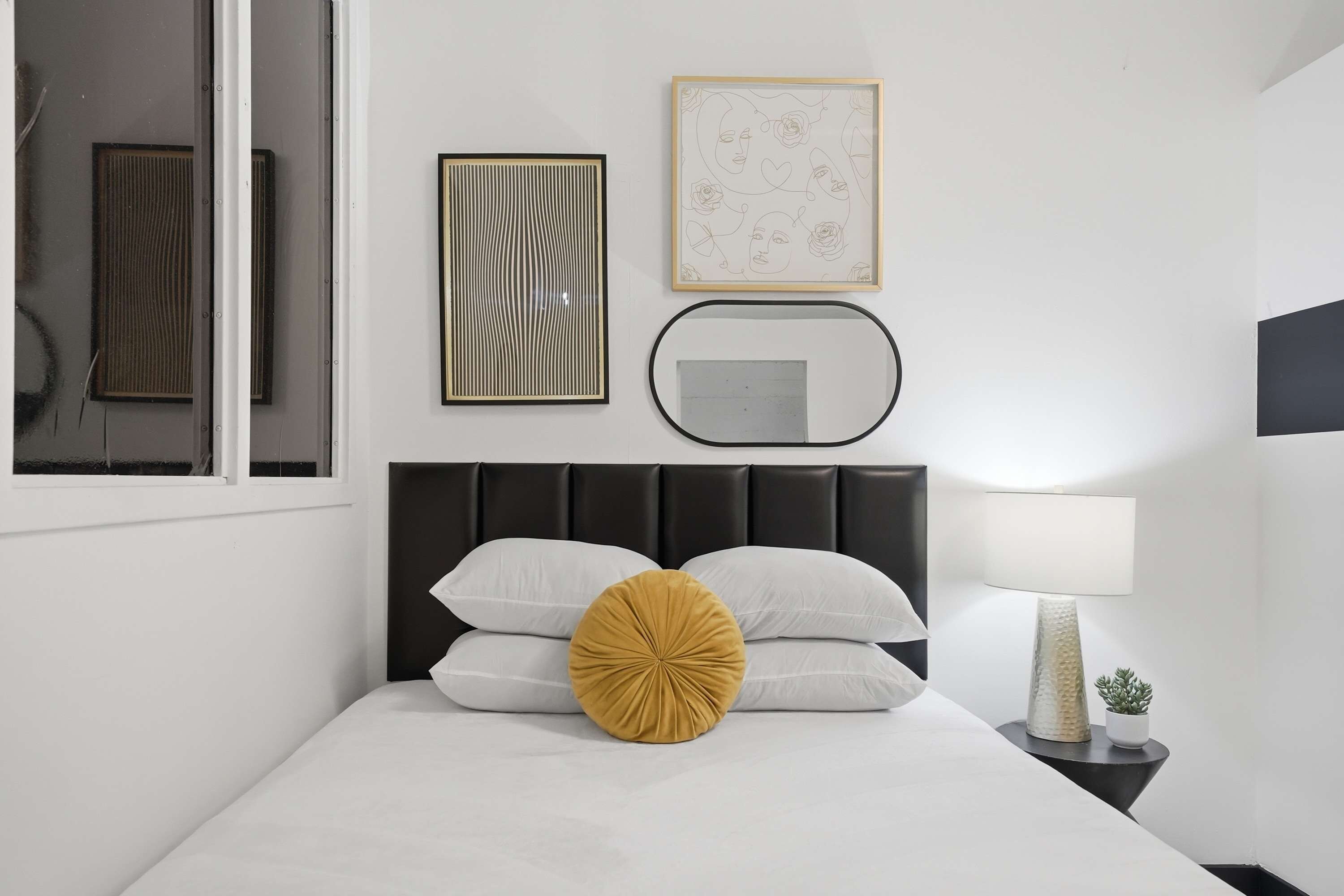 A bedroom with a bed, a bedside lamp, a small potted plant, and framed artwork on the wall.