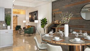 Un comedor moderno con una mesa de madera dispuesta para seis personas, rodeada de sillas blancas. Hay una planta en maceta grande y un jarrón blanco con ramas sobre la mesa.
