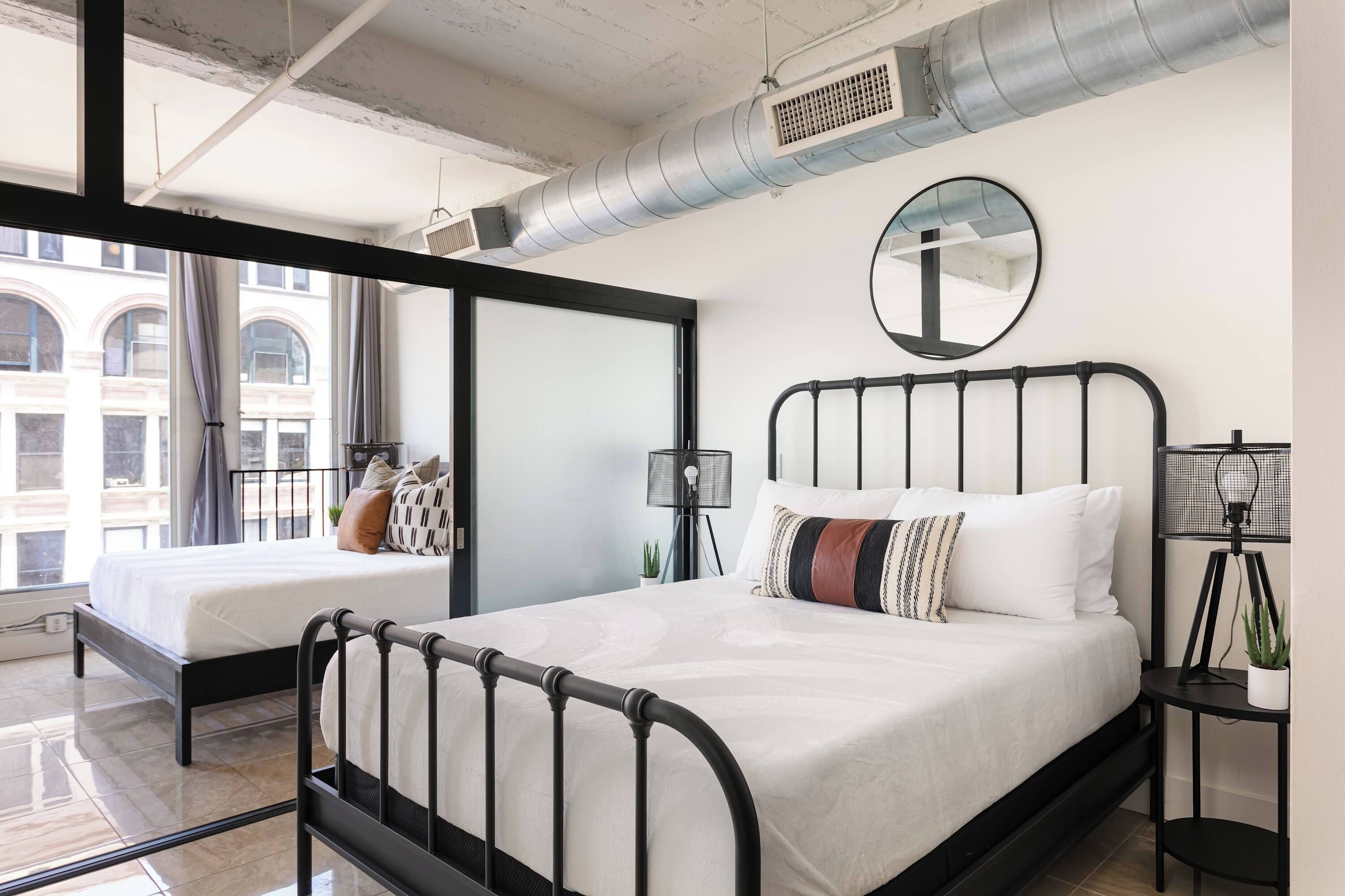Quarto moderno com estrutura de cama em metal preto, roupa de cama branca, um espelho redondo e uma pequena mesa de cabeceira com luminária e planta.