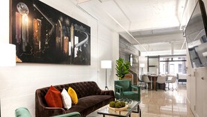Un salón moderno con un sofá marrón, sillones verdes, una mesa de centro de cristal y un cuadro abstracto grande en la pared.