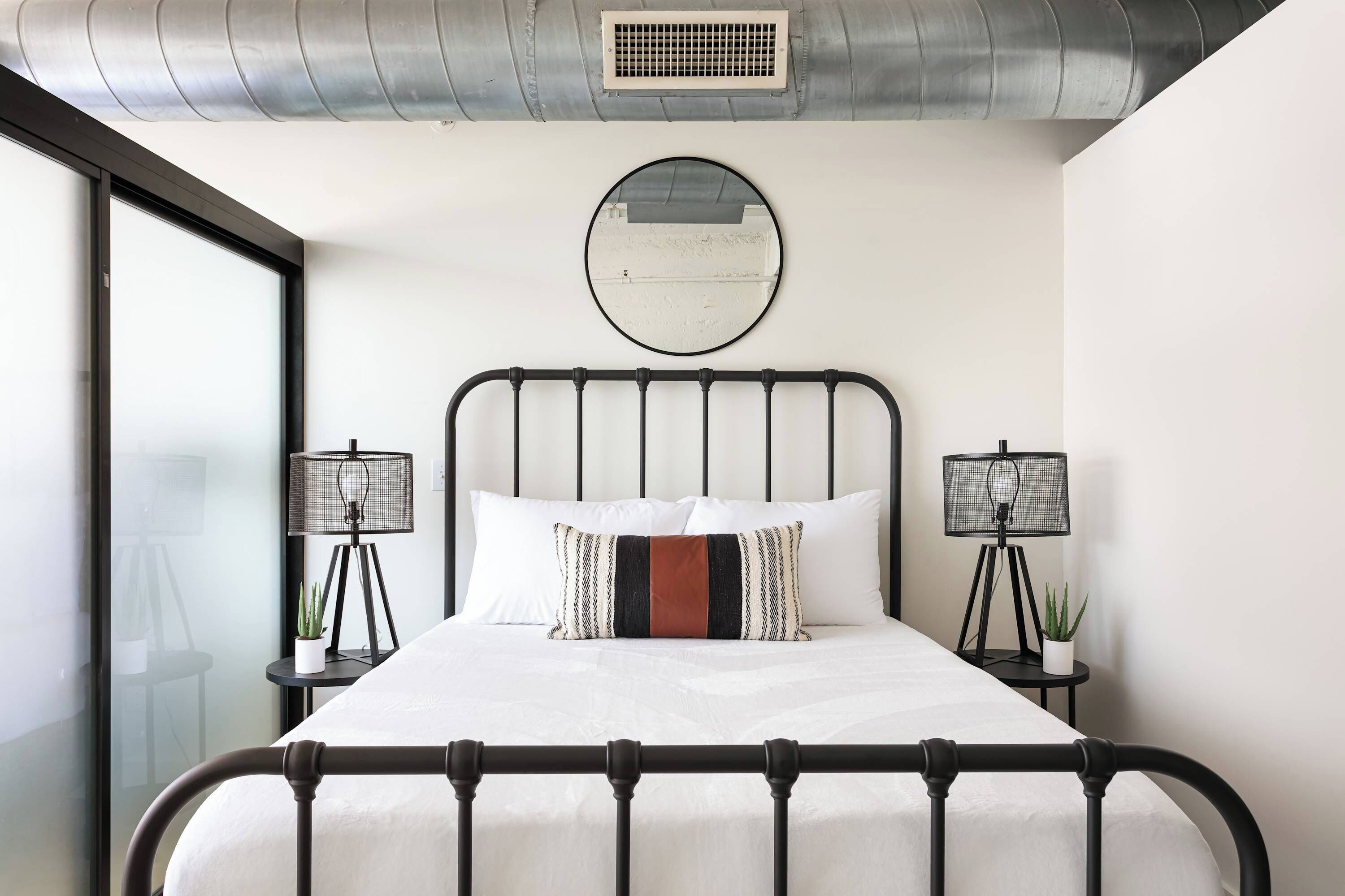 A modern bedroom with a metal bed frame, white bedding, two bedside lamps, and a round mirror on the wall.