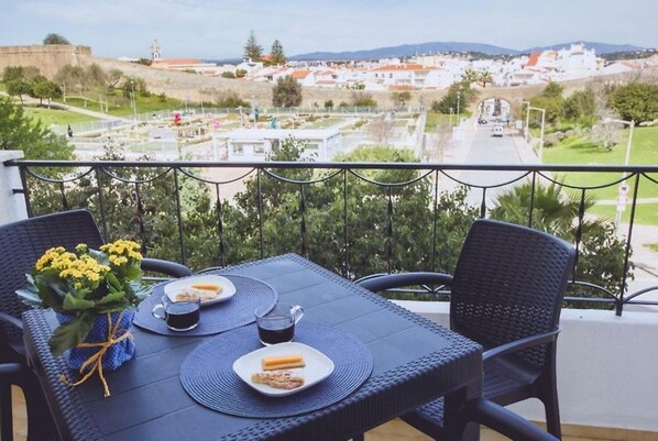 Un balcón con una mesa preparada para dos personas, con vistas a un pueblo con edificios y una colina al fondo.