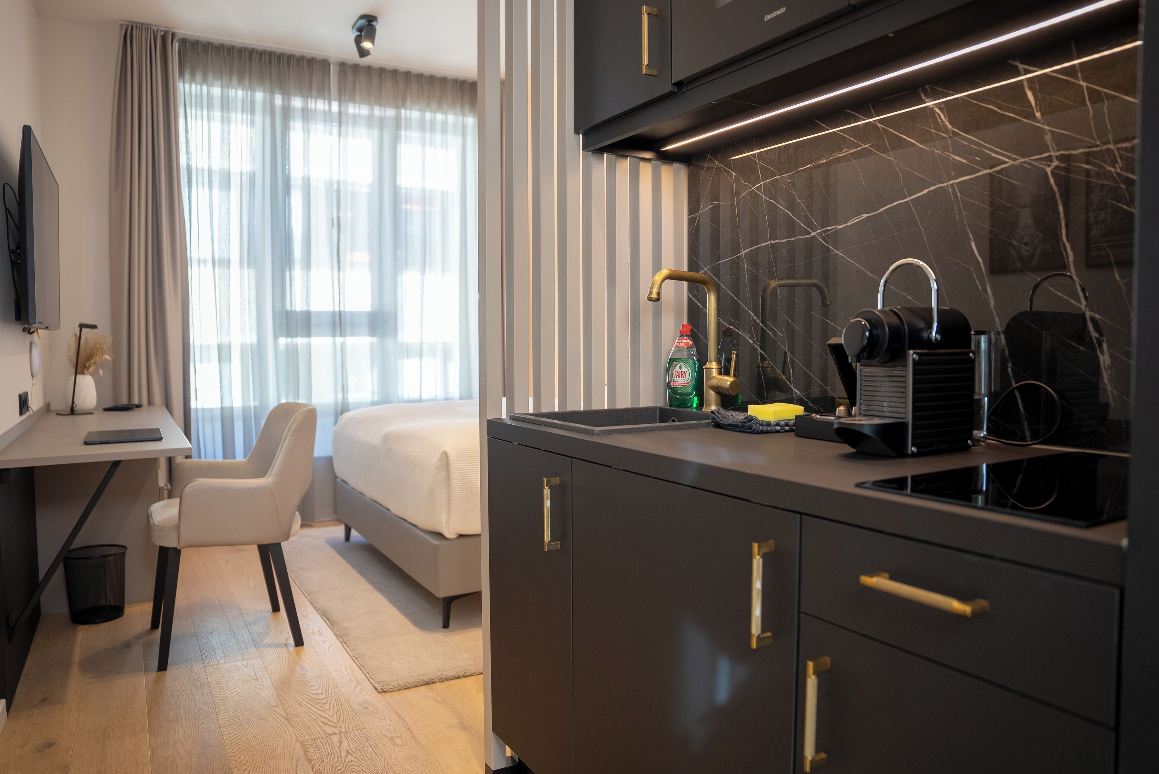 A modern hotel room with a desk, chair, bed, and a kitchen area with a sink and appliances.