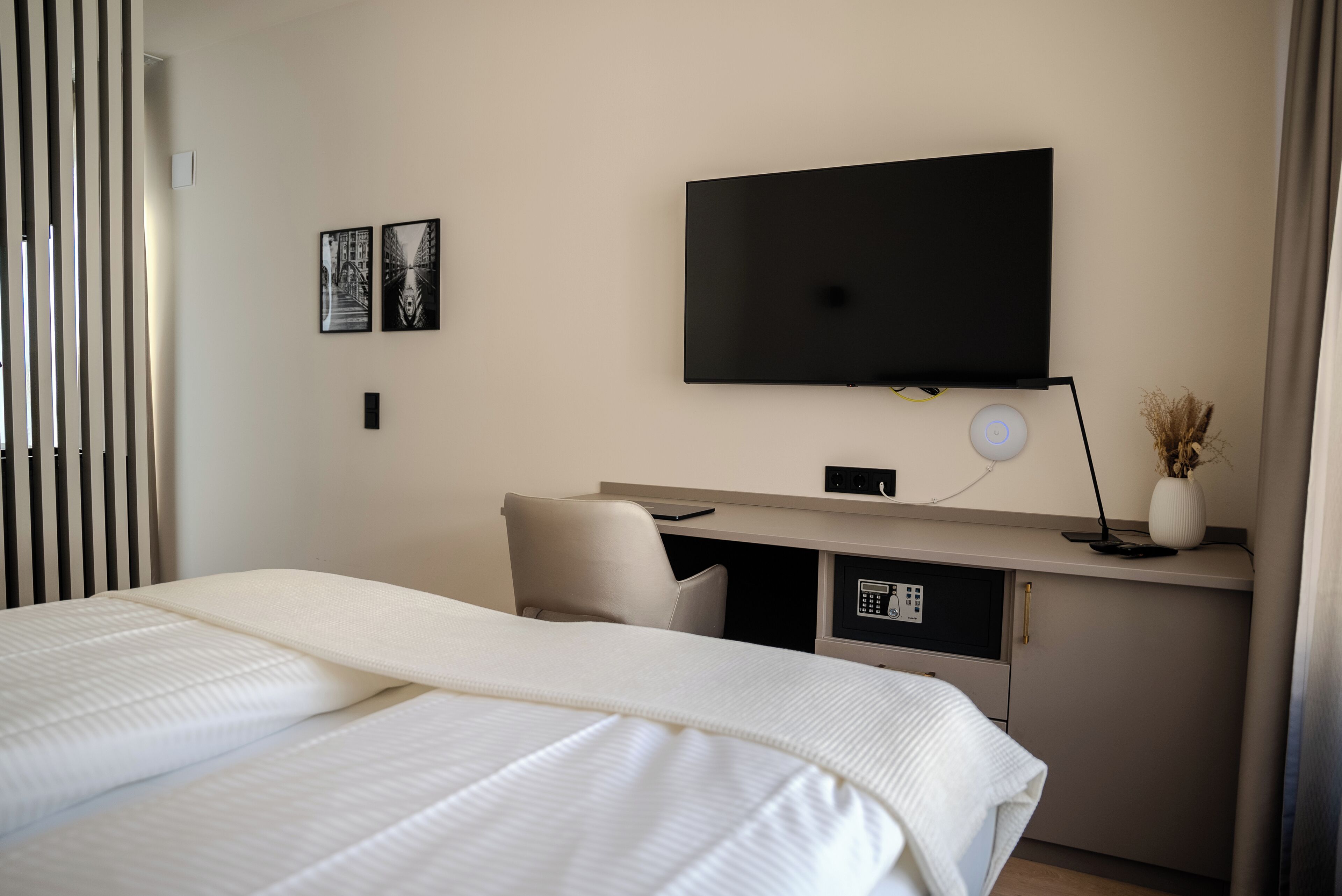 A hotel room with a bed, a desk with a computer, a television, and a wall-mounted control panel.
