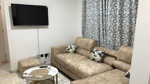 A living room with a beige sofa, two patterned throw pillows, a round coffee table, and a flat-screen TV mounted on the wall. - Kiambu Zone Heights (Nairobi)
