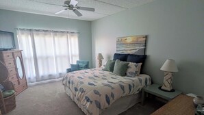 A bedroom with a bed, a ceiling fan, a nightstand with a lamp, a dresser, and a window with curtains. - Pier Point South Ocean View Condos B-15 (St. Augustine)