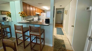 A compact kitchen with wooden cabinets, a central island with bar stools, a refrigerator, and a microwave. - Pier Point South Ocean View Condos B-15 (St. Augustine)