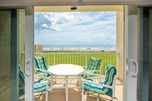 Un balcon avec une table blanche et des chaises à rayures, donnant sur une plage délimitée par une clôture, où l’on aperçoit des personnes au loin.