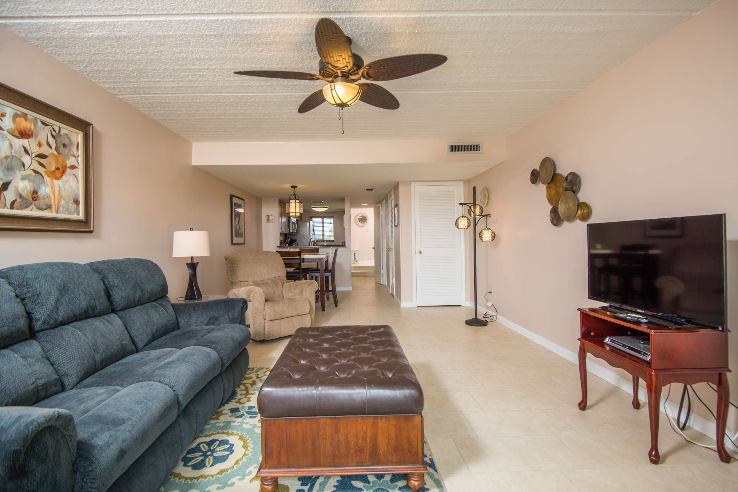 A living room with a sofa, ottoman, TV stand, ceiling fan, and artwork on the wall.