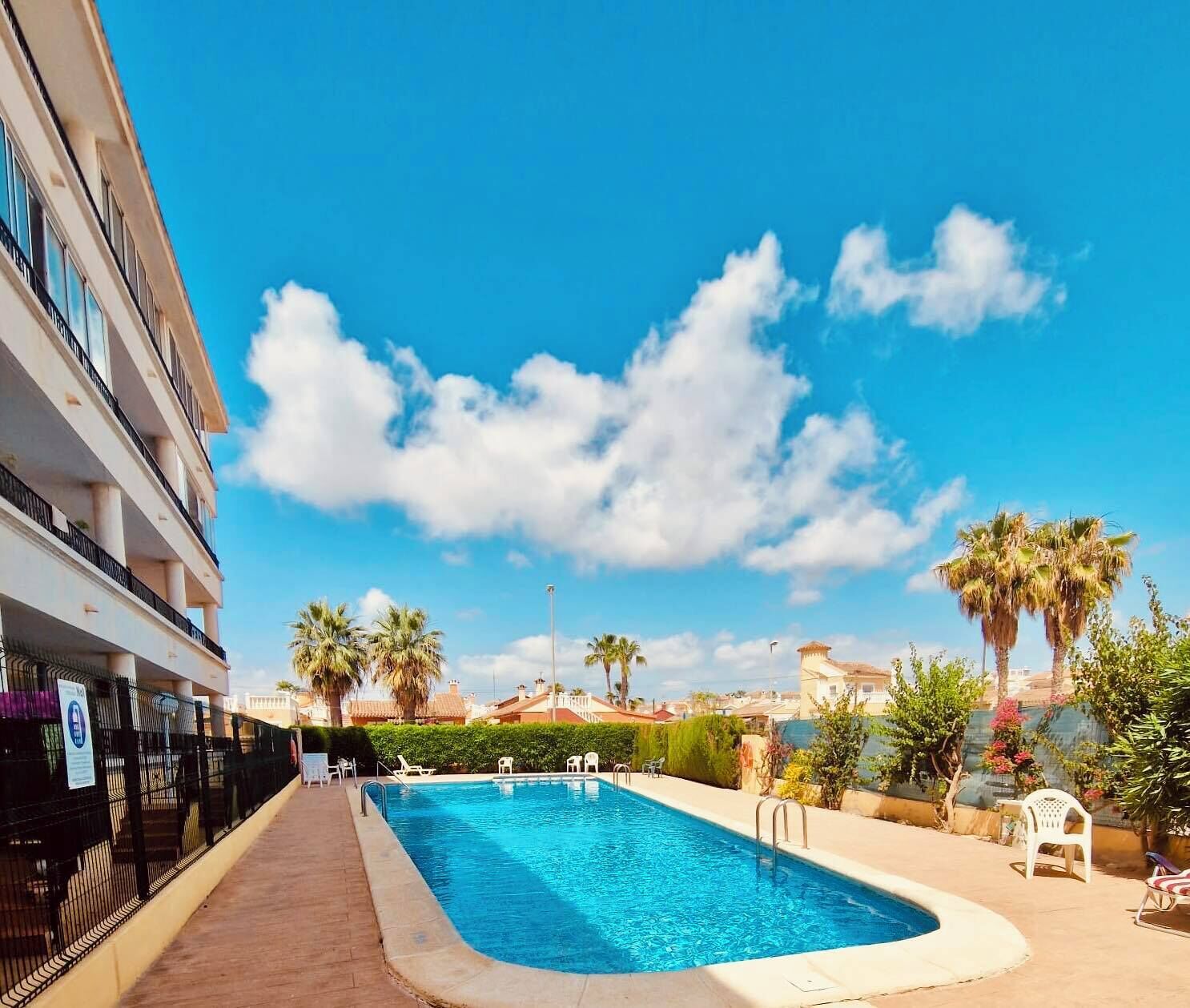A swimming pool with a clear blue sky and clouds, surrounded by palm trees and a building with balconies.