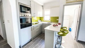 Ett modernt kök med vita skåp, en svart ugn, en grön köksö med barstolar och ett fönster som släpper in naturligt ljus.