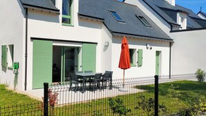 Een tweelaags huis met een groene deur, een terras met een tafel en stoelen, en een rode parasol.