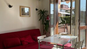 A red sofa, a glass table with chairs, and a potted plant in a room with a sliding glass door to an outdoor area. - 3V Apartments UGO WITH POOL (Bardolino)