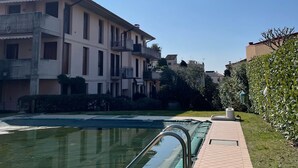A swimming pool with a tiled deck and metal railings, surrounded by a well-maintained hedge and a row of buildings with balconies. - 3V Apartments UGO WITH POOL (Bardolino)