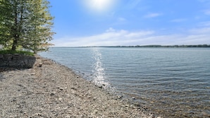 A pebbled beach with a stone wall and a tree, leading to calm water under a clear sky.