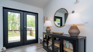 A modern interior with a dark wooden console table, two lamps, a large mirror, and a sliding glass door leading to a deck. - New Buffalo With Hot Tub and Association Beach! (New Buffalo)