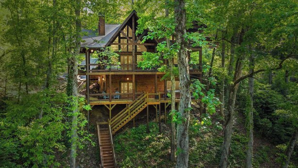 A multi-level wooden cabin with a staircase leading to a deck, surrounded by dense forest.