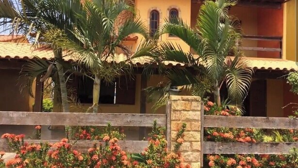 A building with a tiled roof and a stone wall. There are palm trees and flowering plants in front of the building. - Holiday home in Araruama/ Praia Seca. Located in the Palm Beach condominium (Praia Seca)