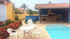 A swimming pool area with a patio table and chairs, surrounded by a fence and a building with a tiled roof. - Holiday home in Araruama/ Praia Seca. Located in the Palm Beach condominium (Praia Seca)