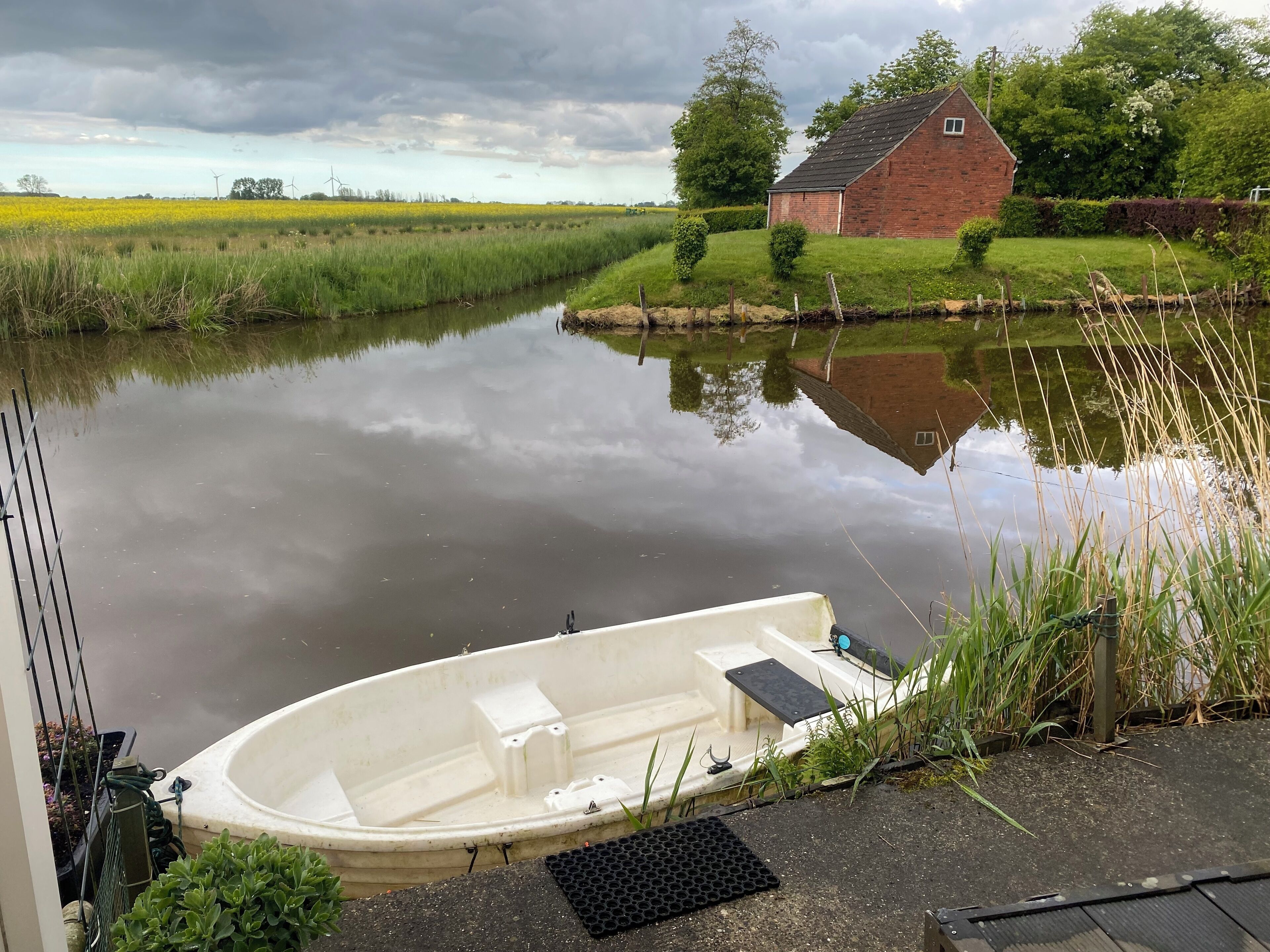 Ein ruhiger Fluss mit einem vertäuten Boot am Ufer, eine Wiese und ein Backsteinhaus im Hintergrund.