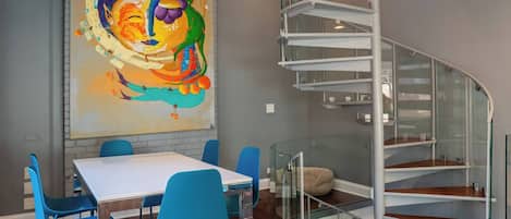 A modern dining area with a white table and blue chairs, a large abstract painting on the wall, and a spiral staircase with glass railings.