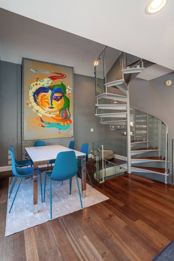 A modern dining area with a white table and blue chairs, a large abstract painting on the wall, and a spiral staircase with glass railings.