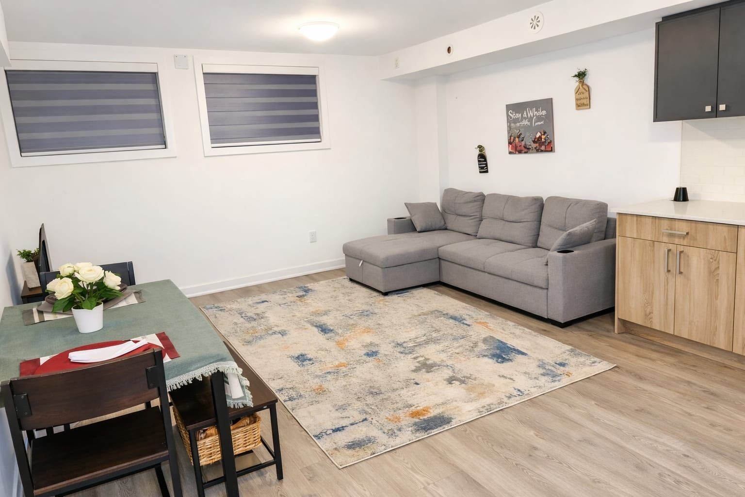 A modern living room with a grey sectional sofa, a dining table with chairs, a patterned rug, and a kitchen area with wooden cabinets.